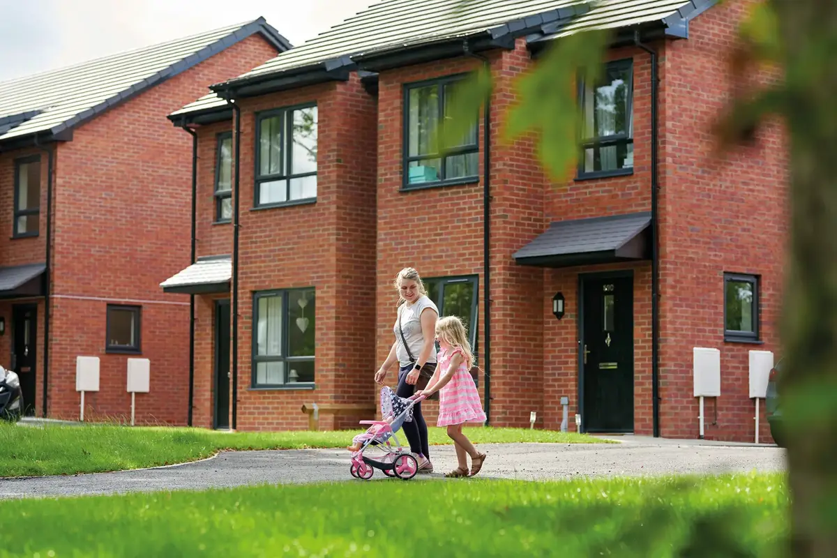 A mum pushing a pram past brick terraced homes on a sunny street